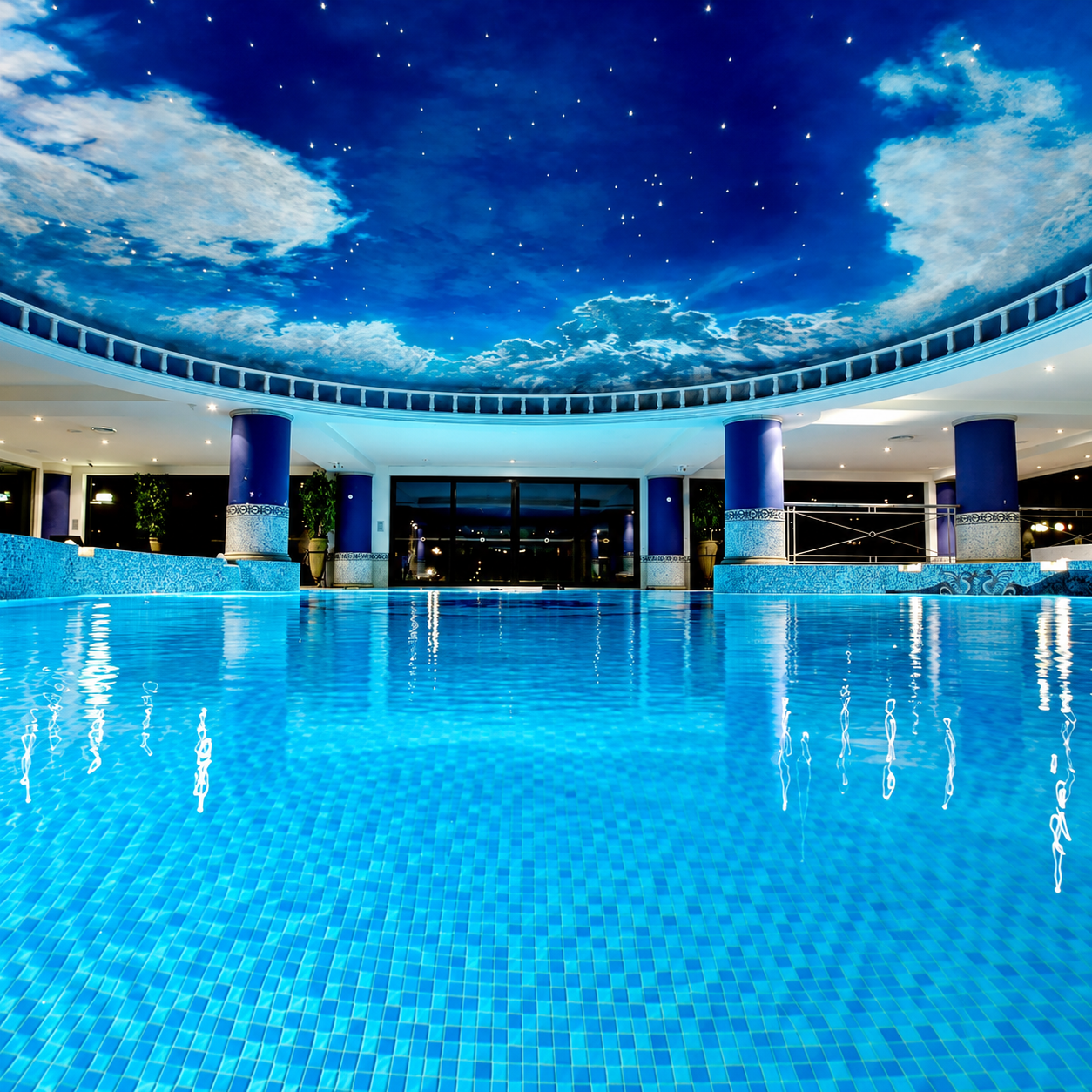 Indoor swimming pool lined with Caribbean blue mosaic tiles beneath a cloud-painted ceiling and decorative columns.
