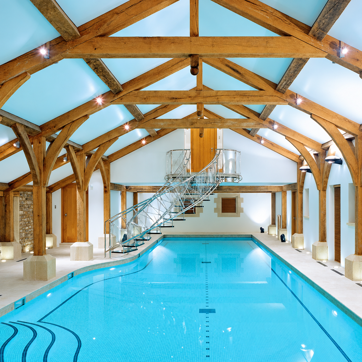 Indoor swimming pool lined with white mosaic tiles beneath turquoise water, with exposed timber beams and a metal staircase.