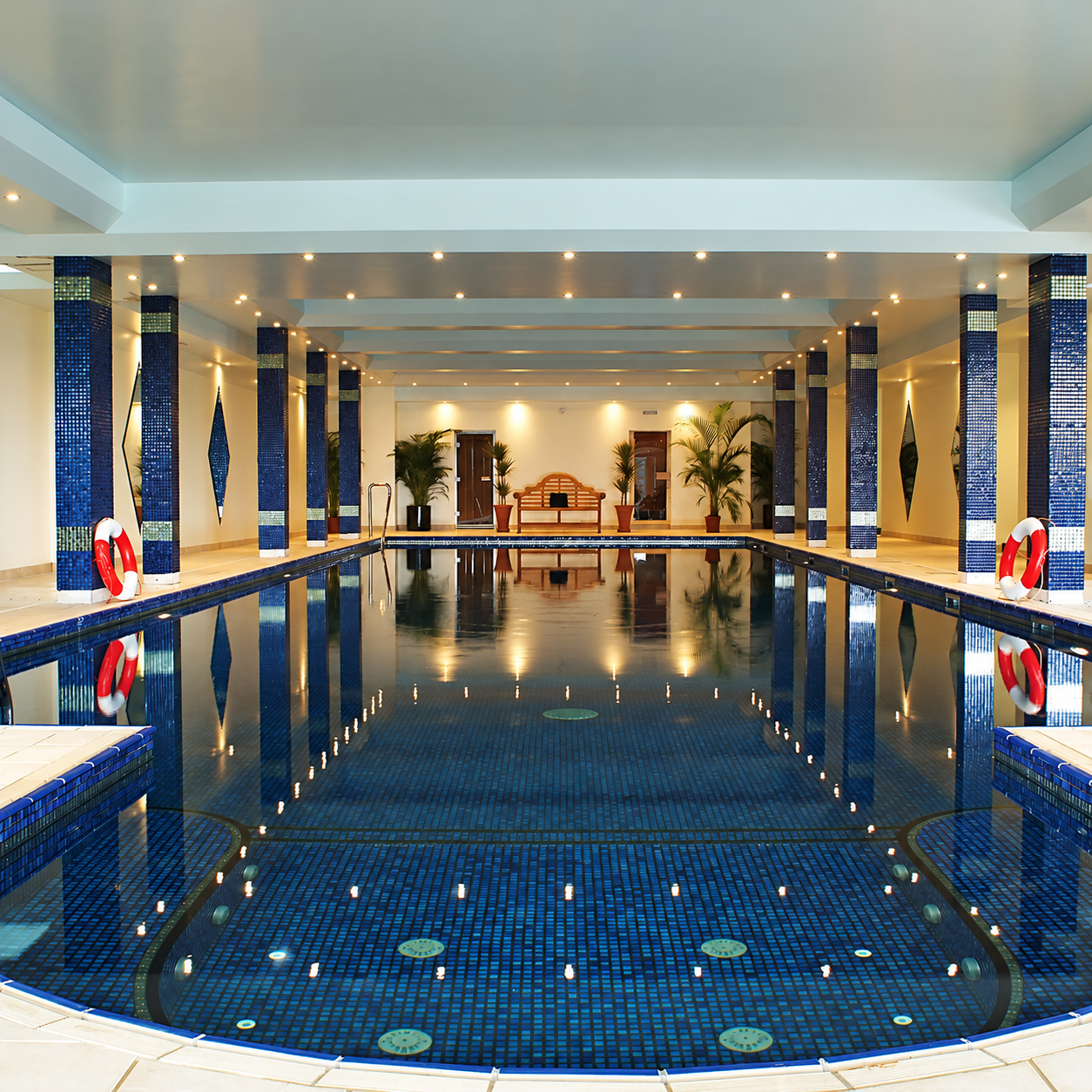 Indoor swimming pool lined with sapphire blue mosaic tiles, with matching tiled columns and warm ceiling lights reflected in the water.