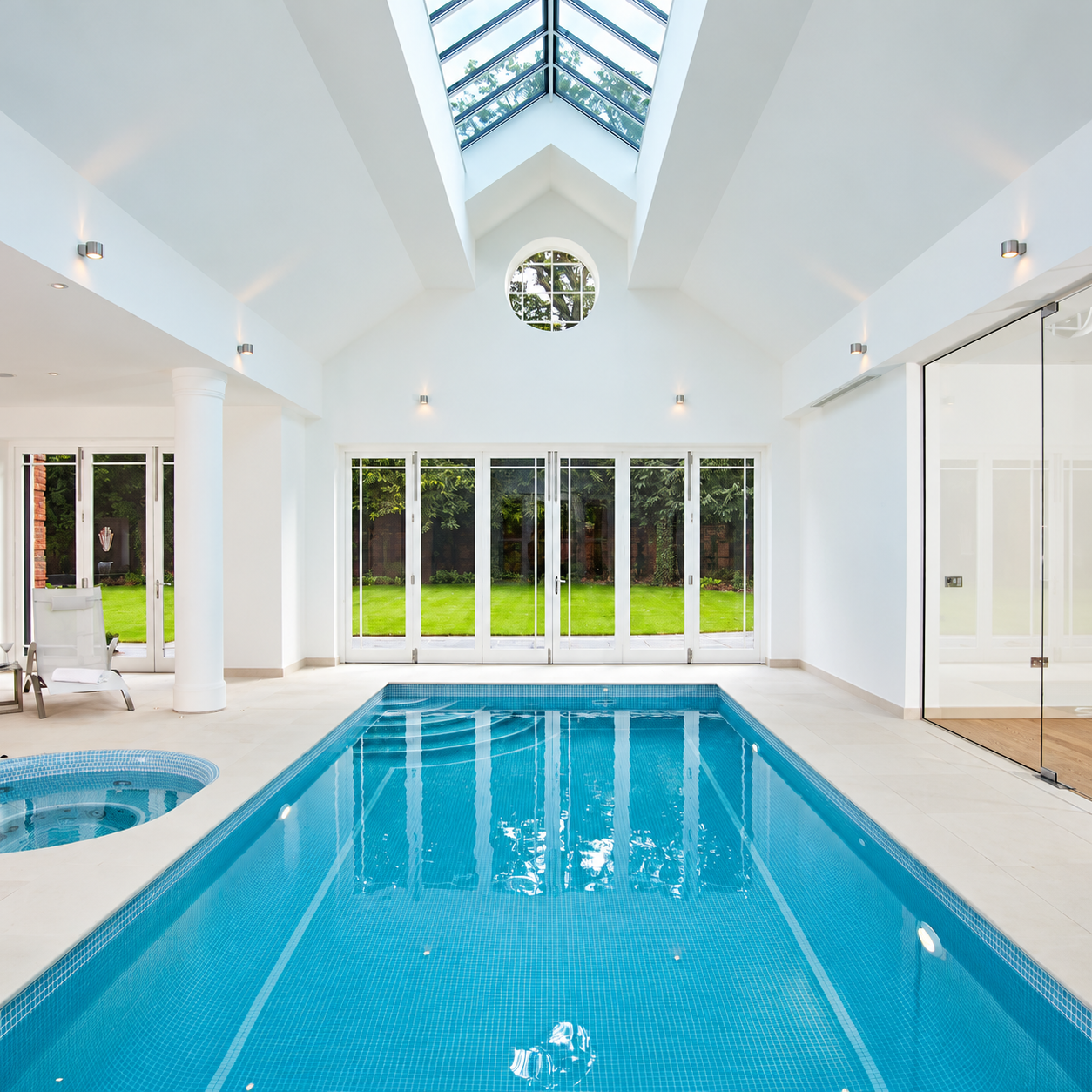 Indoor swimming pool and circular spa lined with sky blue mosaic tiles beneath a skylight, with white walls and garden-facing doors.