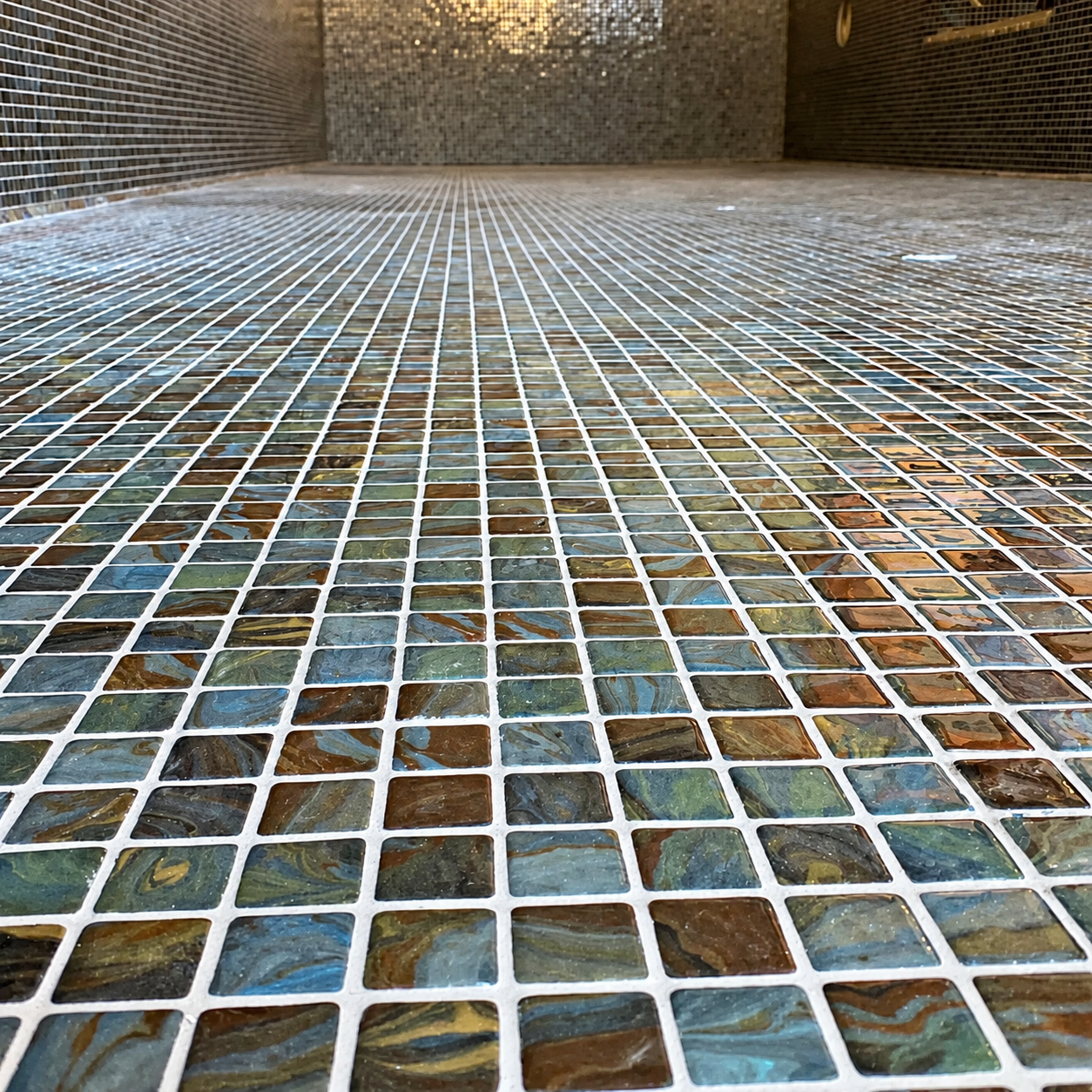 Low-angle view across a mosaic-lined swimming pool tank, showing glossy multicolour squares in a continuous grid.