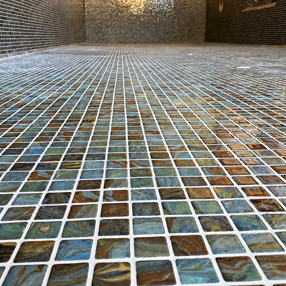 Low-angle view across a mosaic-lined swimming pool tank, showing glossy multicolour squares in a continuous grid.