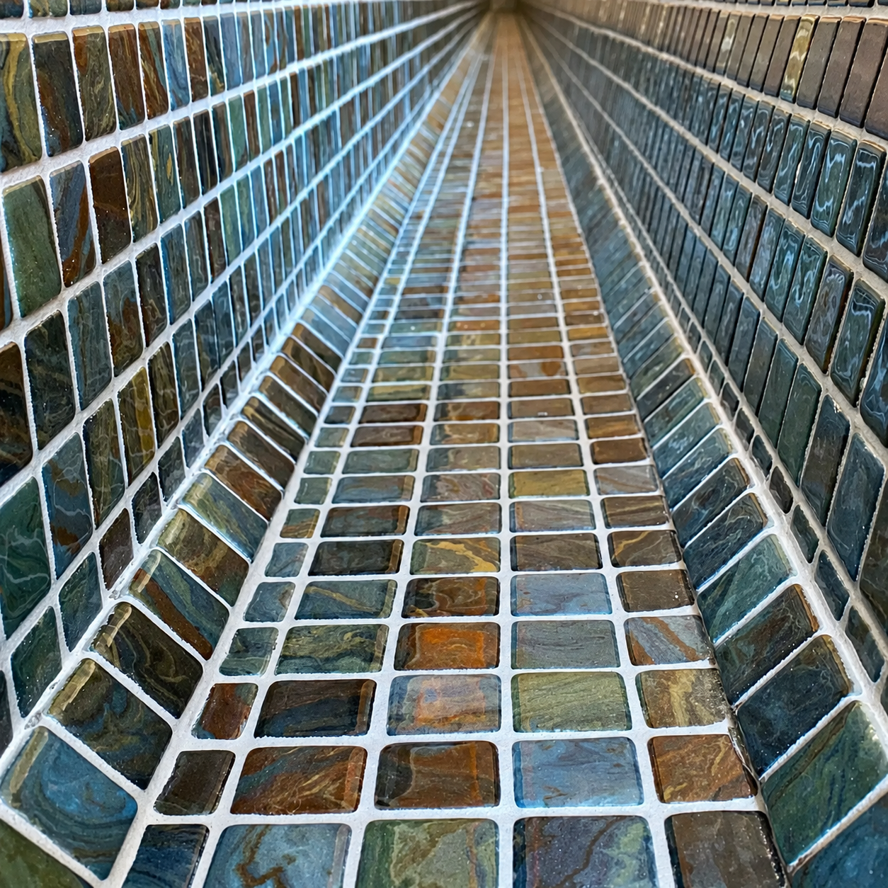 Karma Aquarela glass mosaics lining a narrow pool channel, with blue-green and earthy swirls framed by pale grout.