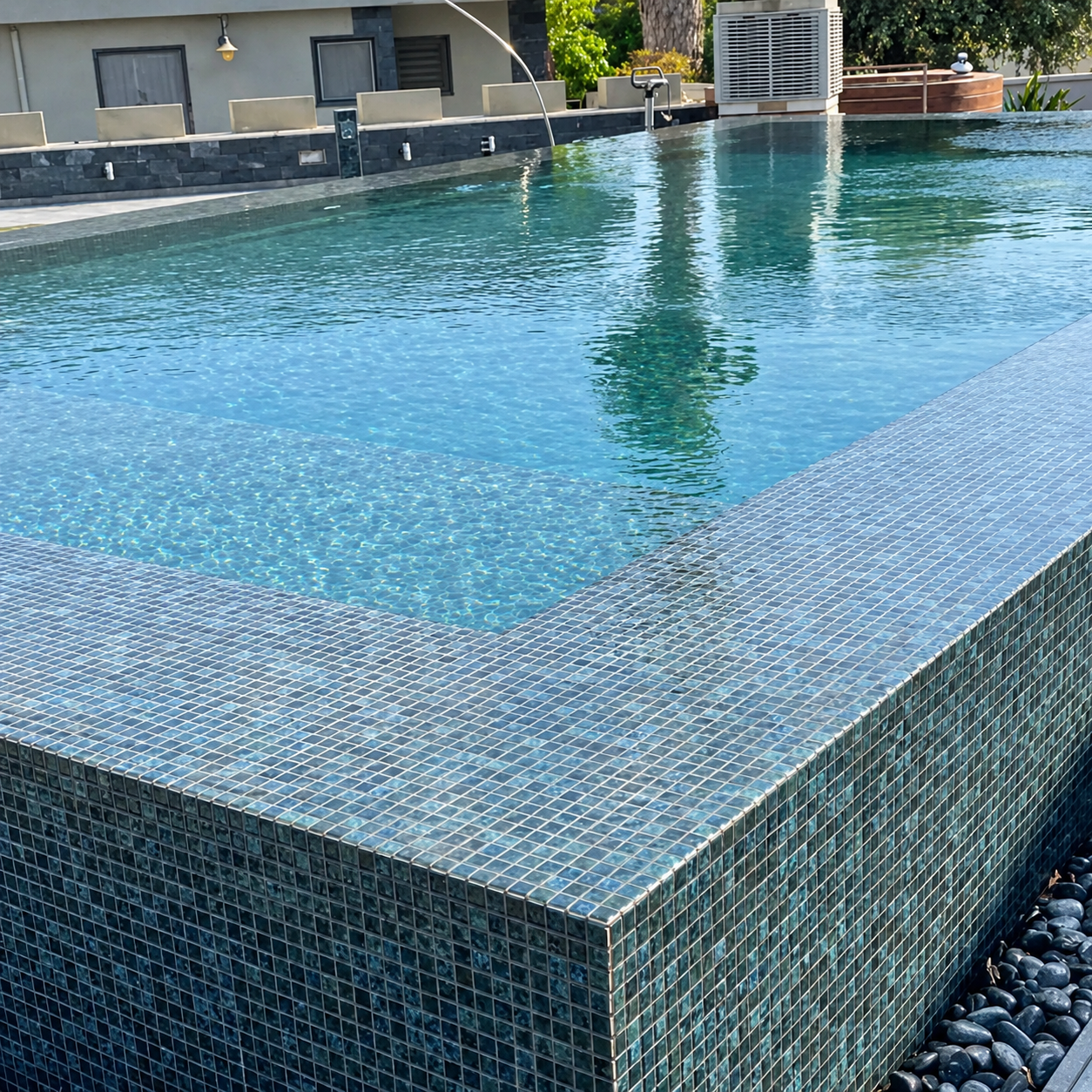 Outdoor swimming pool clad in Karma Blue Moon glass mosaics, with a tiled corner and dark pebble border.