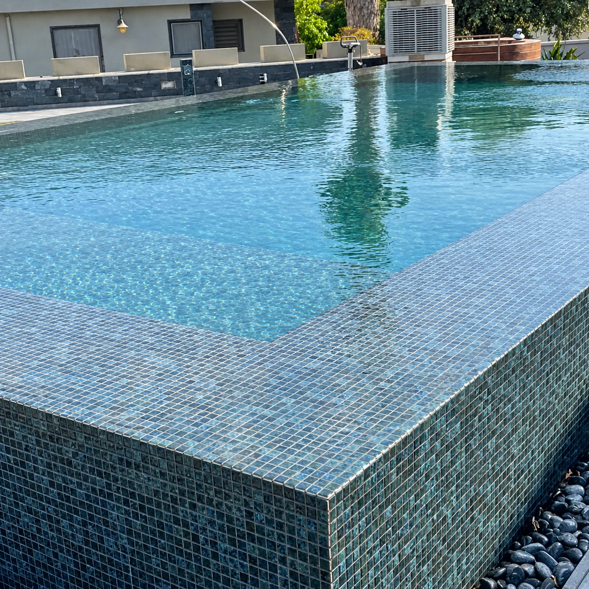 Outdoor swimming pool clad in Karma Blue Moon glass mosaics, with a tiled corner and dark pebble border.
