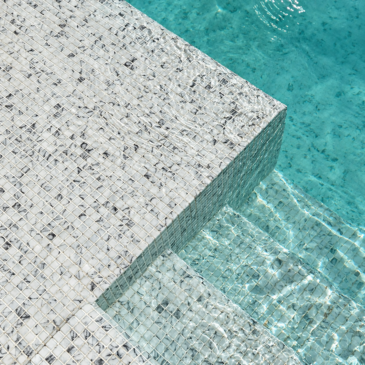 Close-up of Karma Calacatta white and grey glass mosaics on submerged pool steps beneath rippling water.