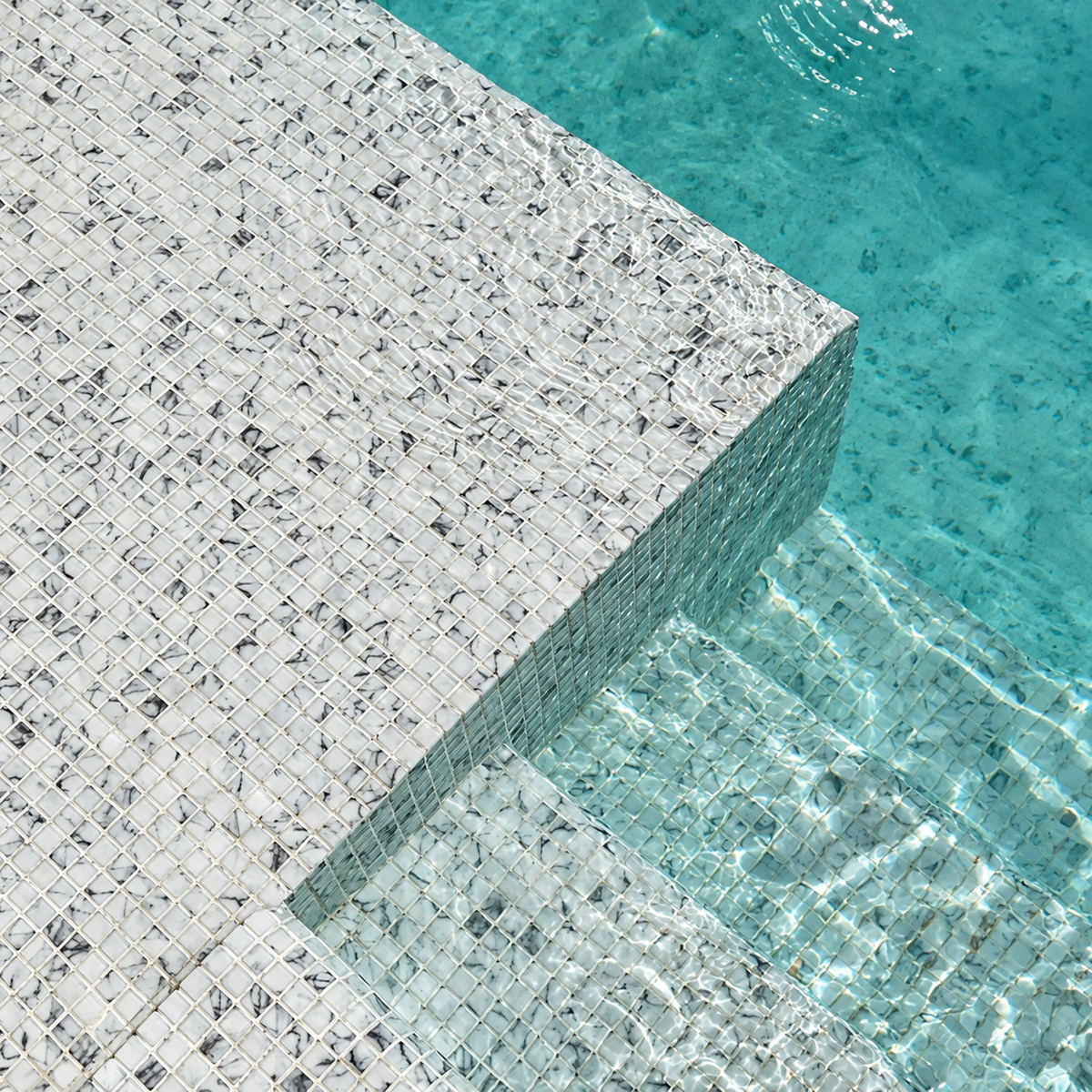 Close-up of Karma Calacatta white and grey glass mosaics on submerged pool steps beneath rippling water.