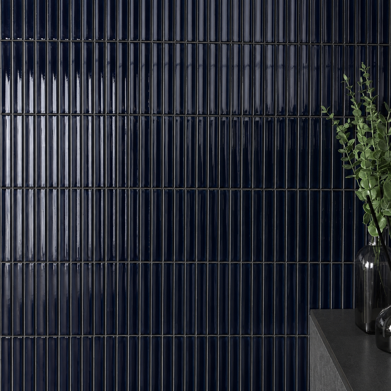 Midnight navy gloss KitKat ceramic mosaic feature wall with slim vertical 20 x 145 mm tiles, 2 mm grout joints, reflective glazed finish, dark stone ledge and black glass vase décor.