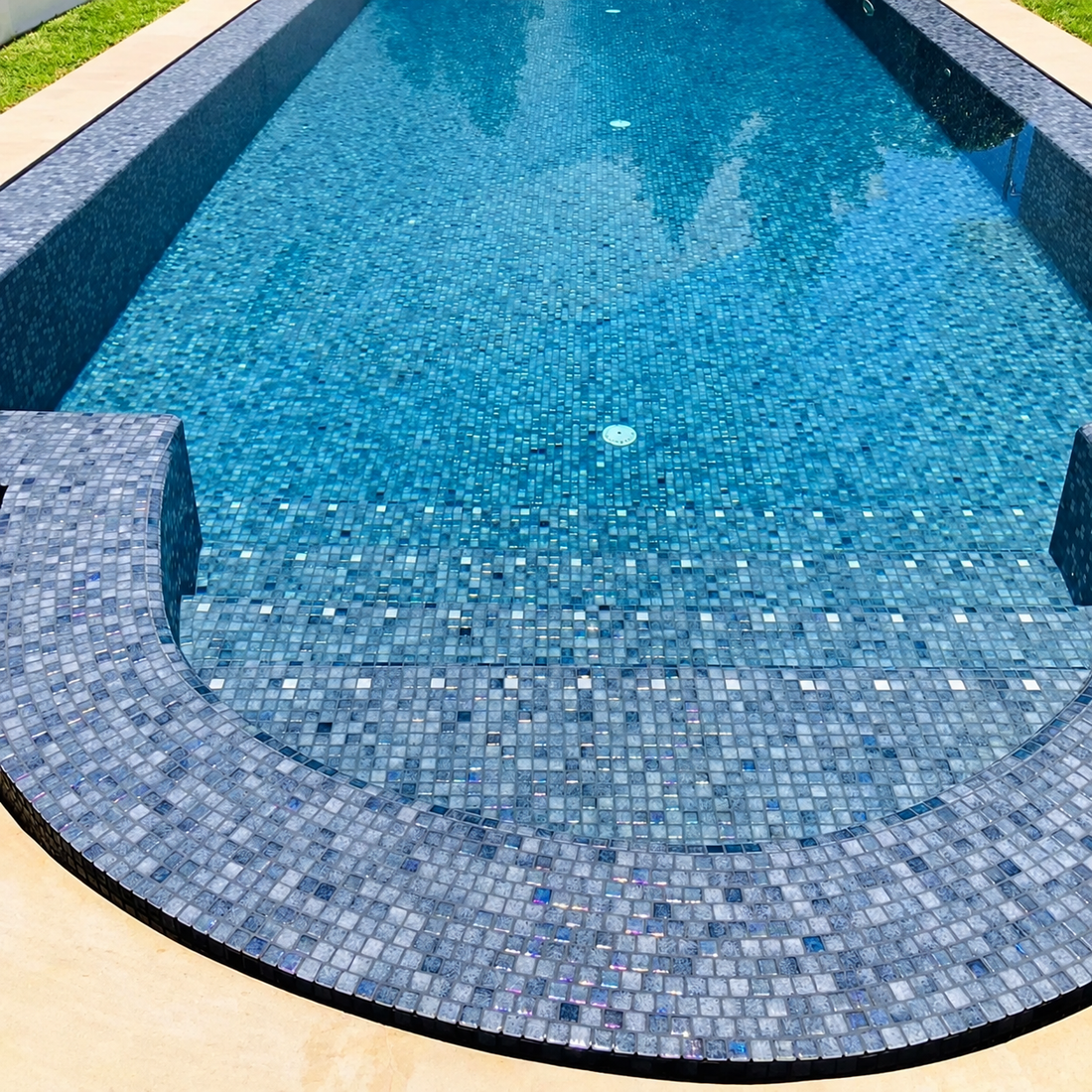 Niebla Mix-Iris Ter grey-purple glass mosaics lining a curved swimming pool edge beneath rippling water.