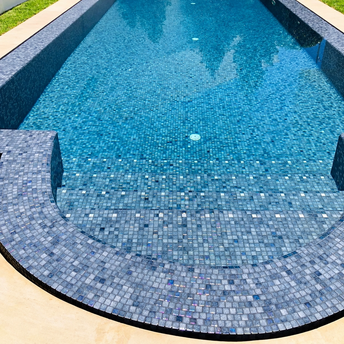 Niebla Mix-Iris Ter grey-purple glass mosaics lining a curved swimming pool edge beneath rippling water.