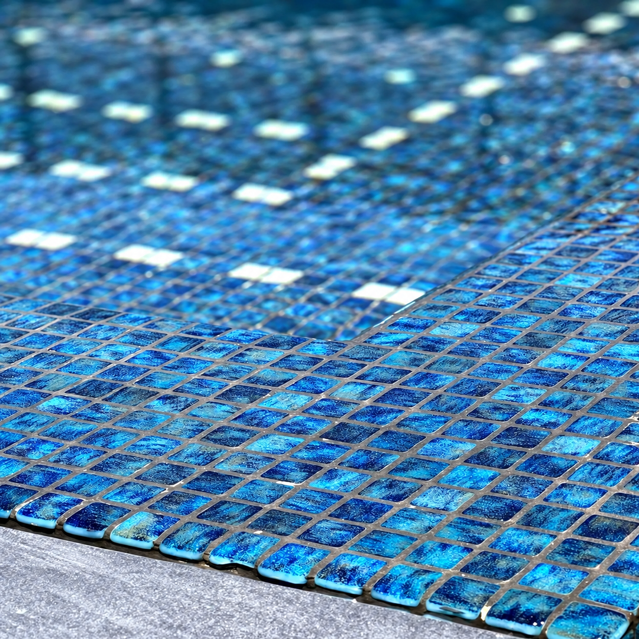 Niebla Paradise Zafiro blue glass mosaics lining a swimming pool edge, with mottled sapphire tones and grey grout.