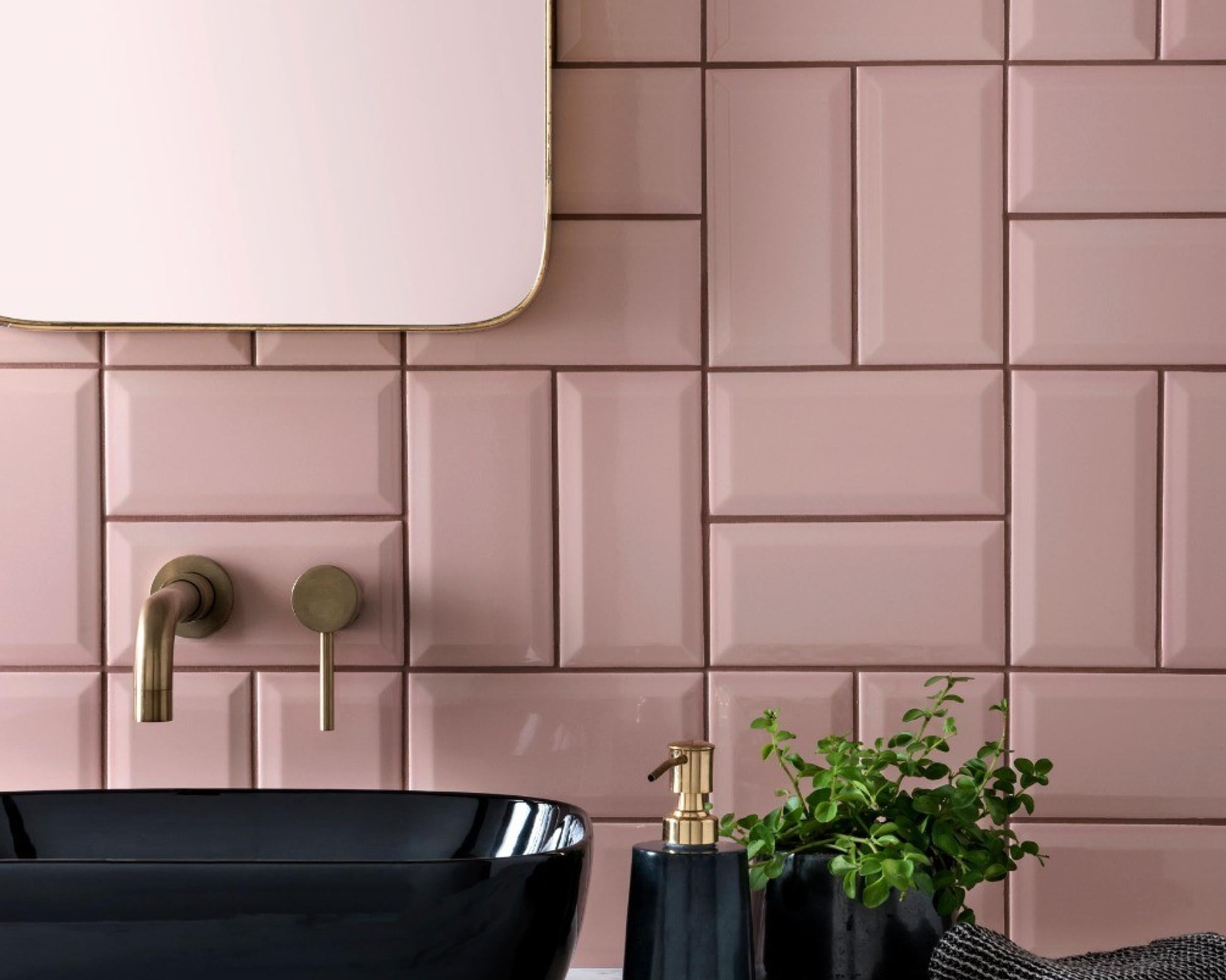 A close-up of a modern bathroom wall featuring glossy, rectangular pink subway tiles arranged in a stacked pattern. A black vessel sink with a matte brass faucet is visible in the foreground, along with a small green potted plant and a black soap dispenser.