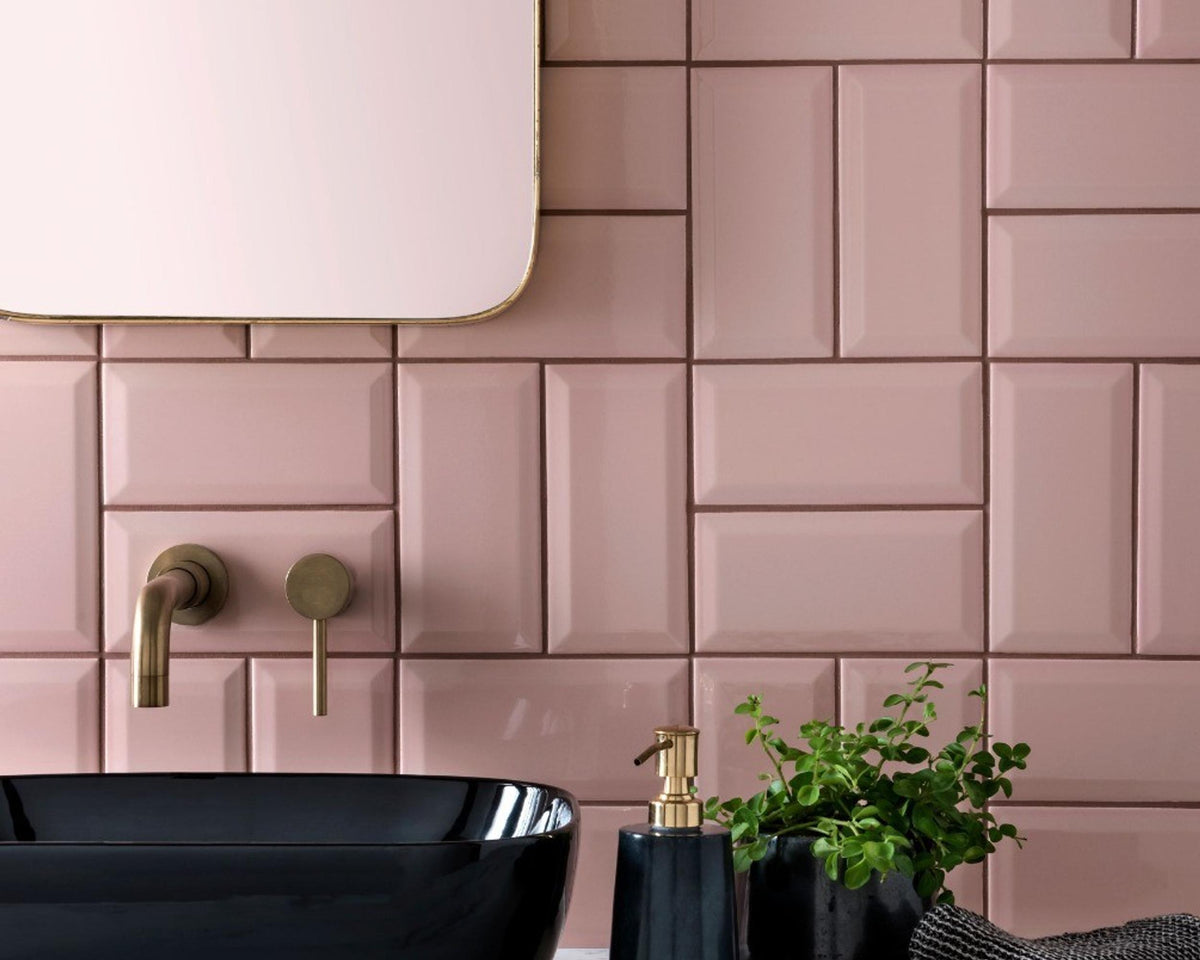 A close-up of a modern bathroom wall featuring glossy, rectangular pink subway tiles arranged in a stacked pattern. A black vessel sink with a matte brass faucet is visible in the foreground, along with a small green potted plant and a black soap dispenser.